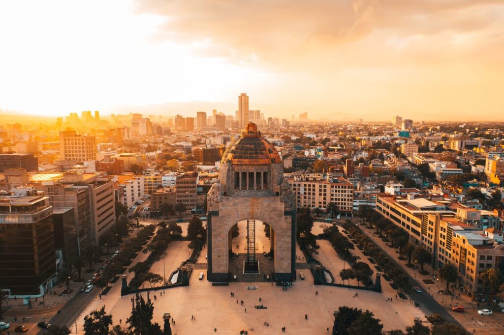 https://www.pexels.com/de-de/foto/stadt-menschen-wahrzeichen-strasse-6942525/ Luftaufnahme von Mexiko-Stadt bei Sonnenuntergang, mit leuchtenden Farben und Gebäuden.