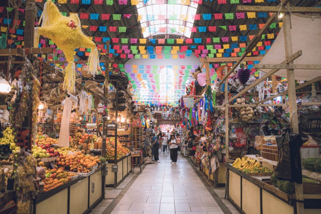 https://www.pexels.com/de-de/foto/lebhaftes-marktgeschehen-in-guanajuato-mexiko-37525346/ Ein Markt mit bunten Fahnen, die von der Decke hängen und eine festliche Atmosphäre schaffen.