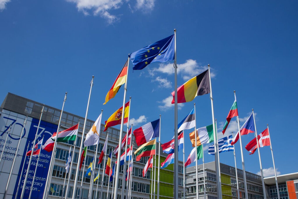 https://www.pexels.com/de-de/foto/fahnen-europaische-union-stadtisch-urban-17178100/ Verschiedene Flaggen der Europäischen Union, vor einem blauen Himmel.