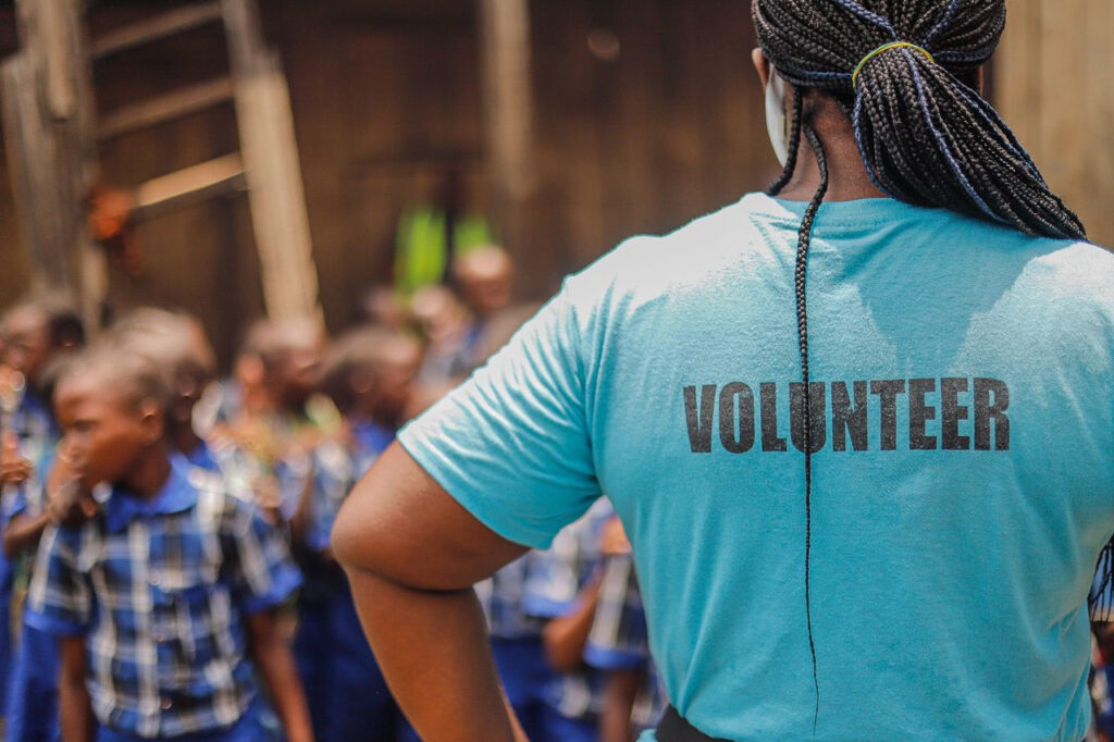 https://www.pexels.com/de-de/foto/frau-dorf-kindheit-gucken-18449721/ Eine Freiwillige in einem blauen Shirt steht gegenüber einer Gruppe von Kindern