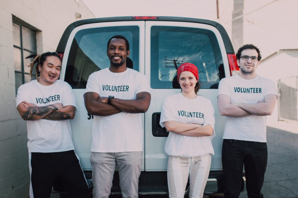 https://www.pexels.com/de-de/foto/liebe-love-menschen-freunde-6647175/ Vier Personen in Freiwilligen-Shirts stehen vor einem Lieferwagen und lächeln in die Kamera.